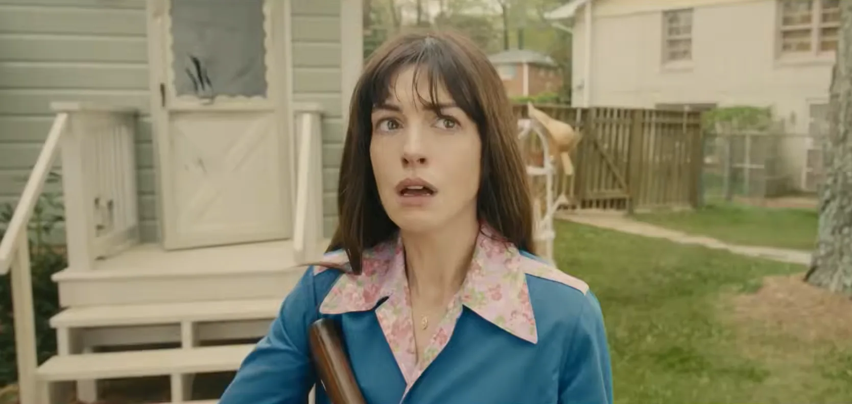 Anne Hathaway, con un arma en la mano y gesto de asombro, frente a las casas de la calle Oak