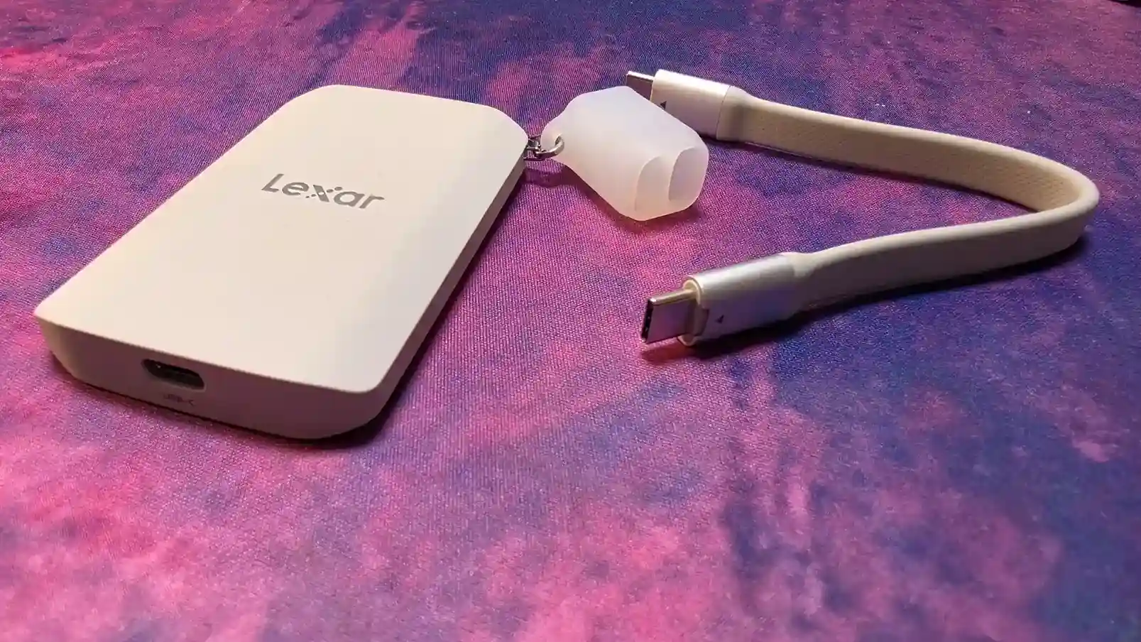 La SSD Lexar ES5 de perfil sobre una tela violeta, con el puerto USB-C a la vista, el organizador de silicona y el cable corto incluido