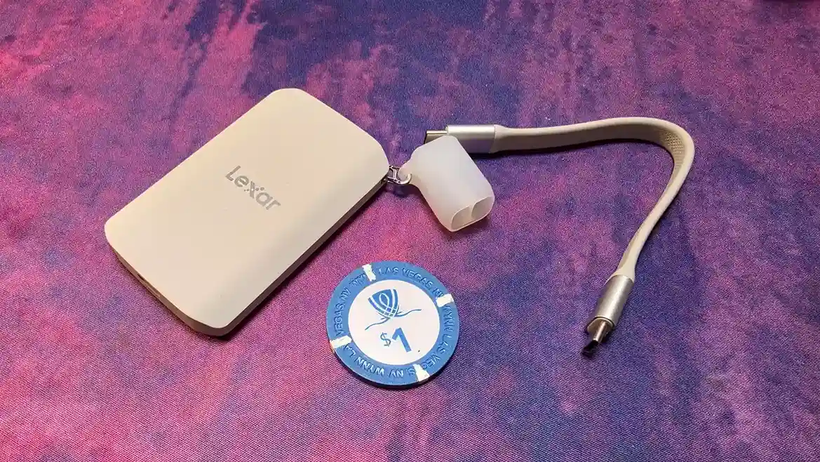 La SSD Lexar ES5 apoyada sobre una tela violeta junto a su cable USB-C y una ficha de casino de un dólar que sirve de referencia de tamaño