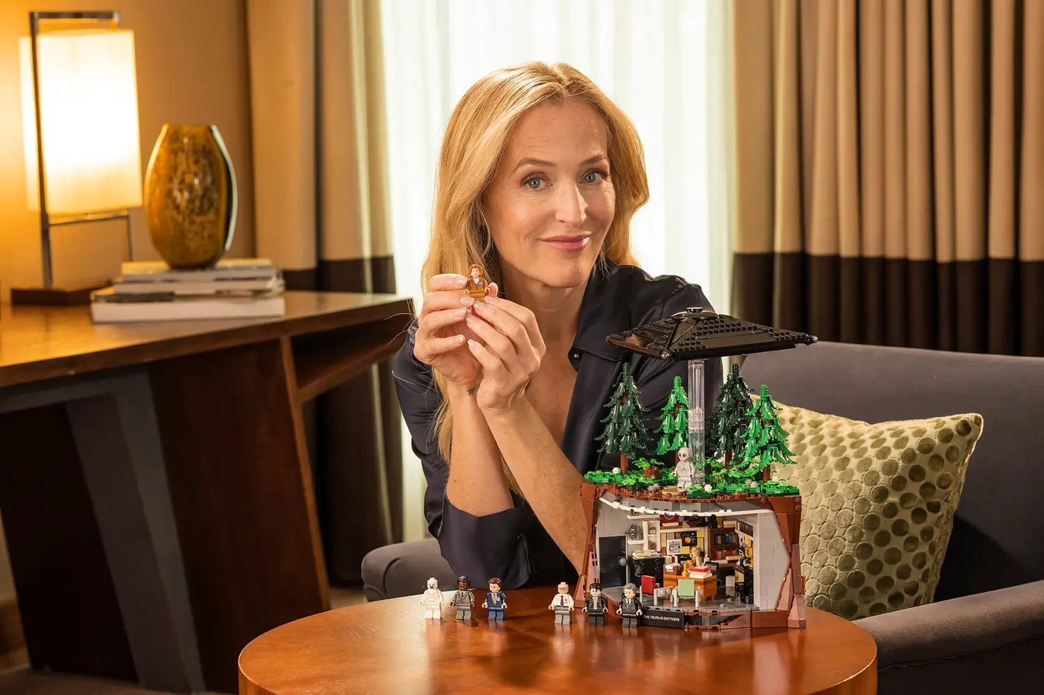 Gillian Anderson sostiene una minifigura de Scully junto al set LEGO X-Files 21369 con la oficina de Mulder y un OVNI