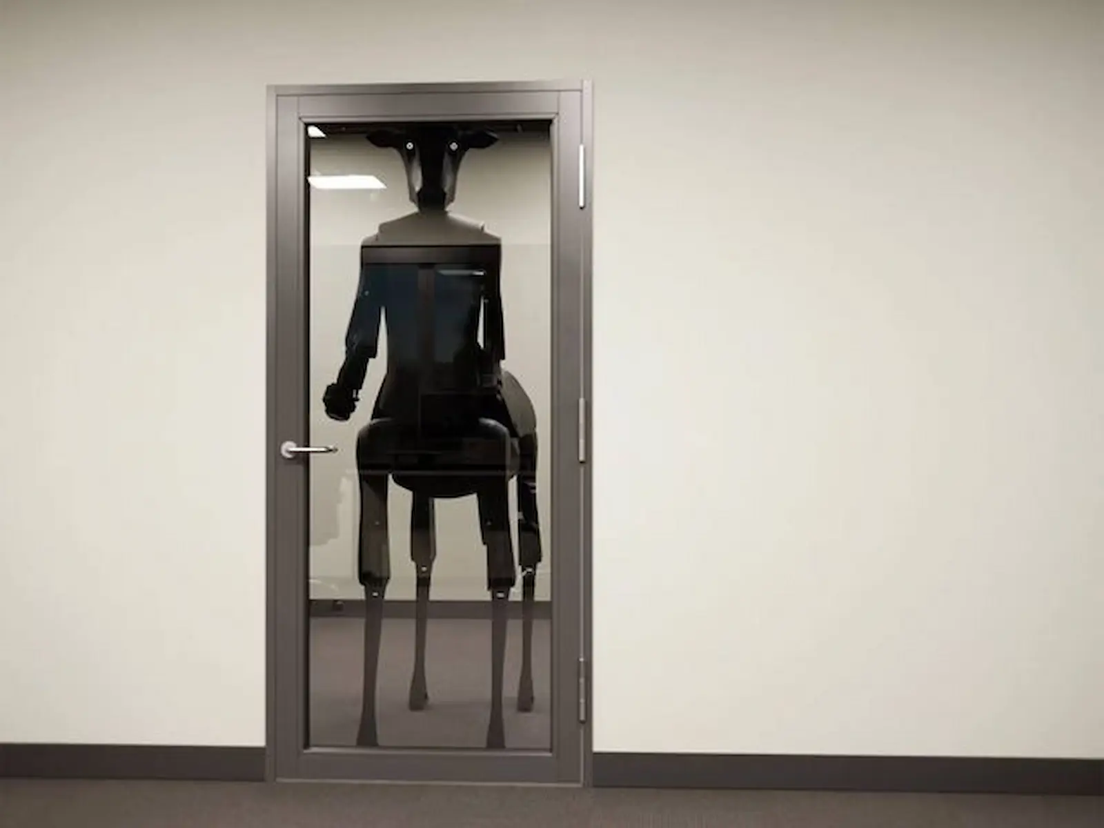 El robot Threehalves de Satyress visto a través de una puerta vidriada cerrada en una oficina, con la cabeza casi al ras del dintel