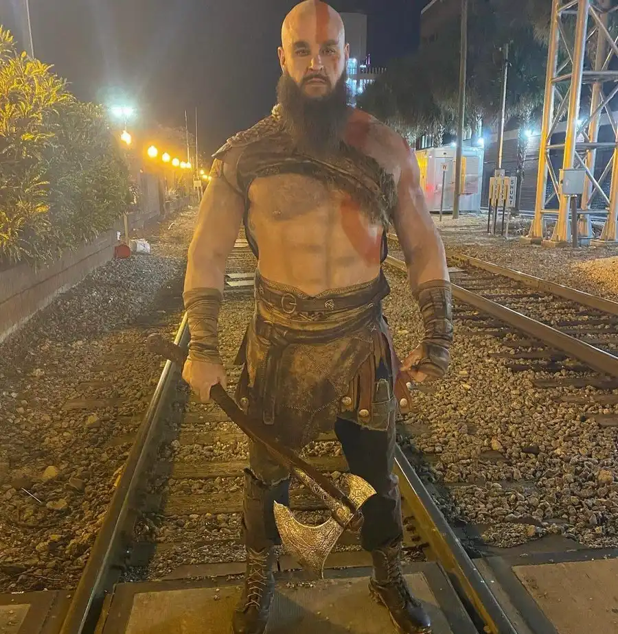 Braun Strowman caracterizado como Kratos, con el torso pintado, el tatuaje rojo y el hacha Leviatán, parado sobre unas vías de tren