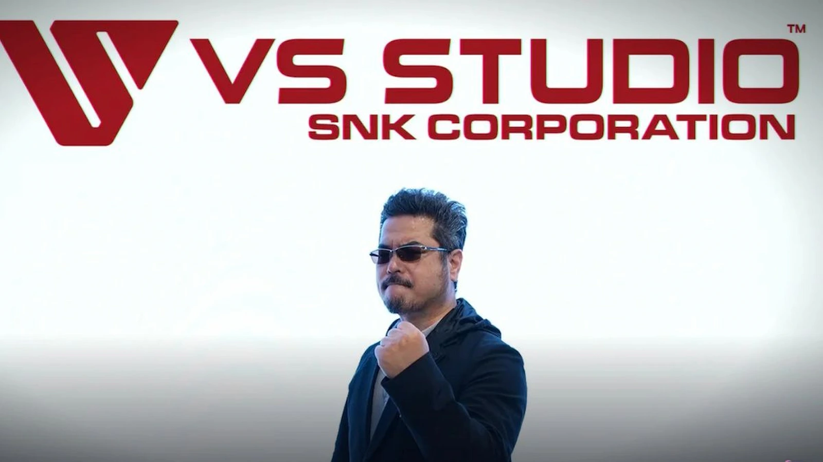 Katsuhiro Harada con el puño en alto debajo del logo de VS Studio, de SNK Corporation