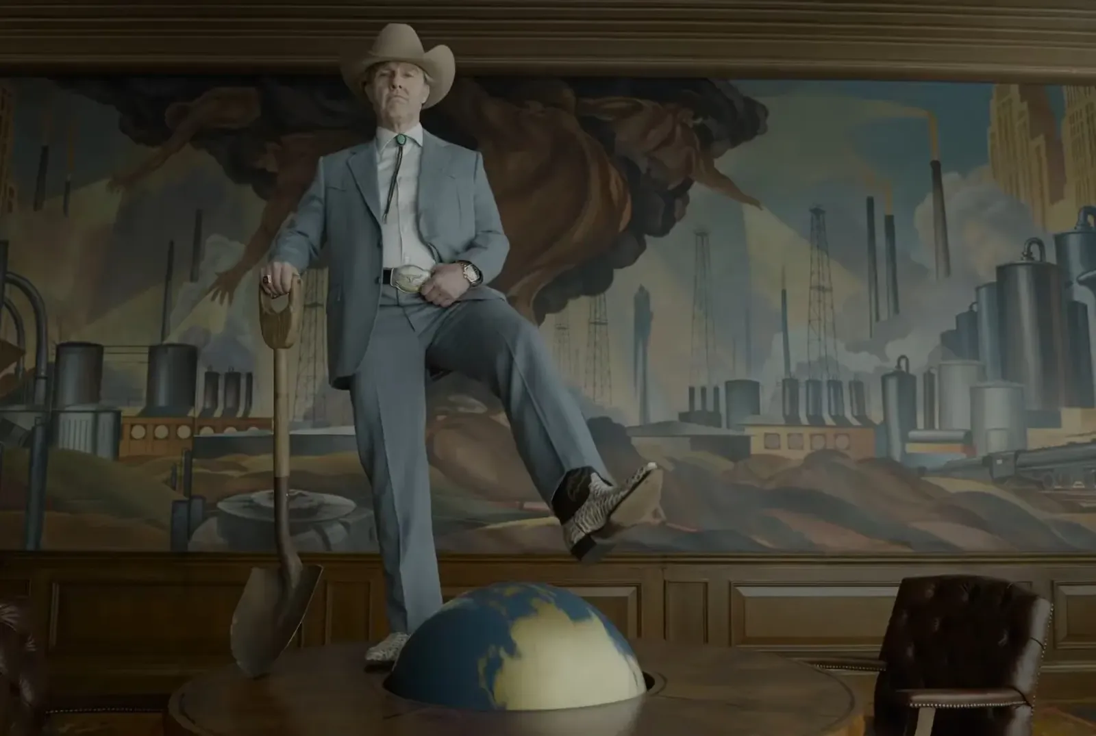Tom Cruise caracterizado como el magnate petrolero Digger Rockwell, con sombrero, una pala en la mano y el pie sobre un globo terráqueo, frente a un mural de torres de petróleo