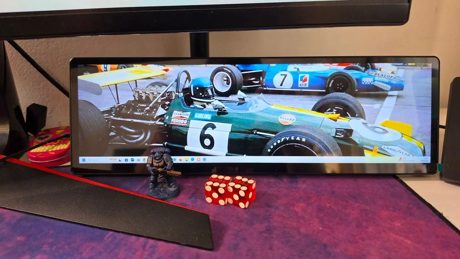 Corsair Xeneon Edge apoyada en su soporte debajo del monitor, usada como segunda pantalla de Windows con un fondo de autos de Fórmula 1 clásicos
