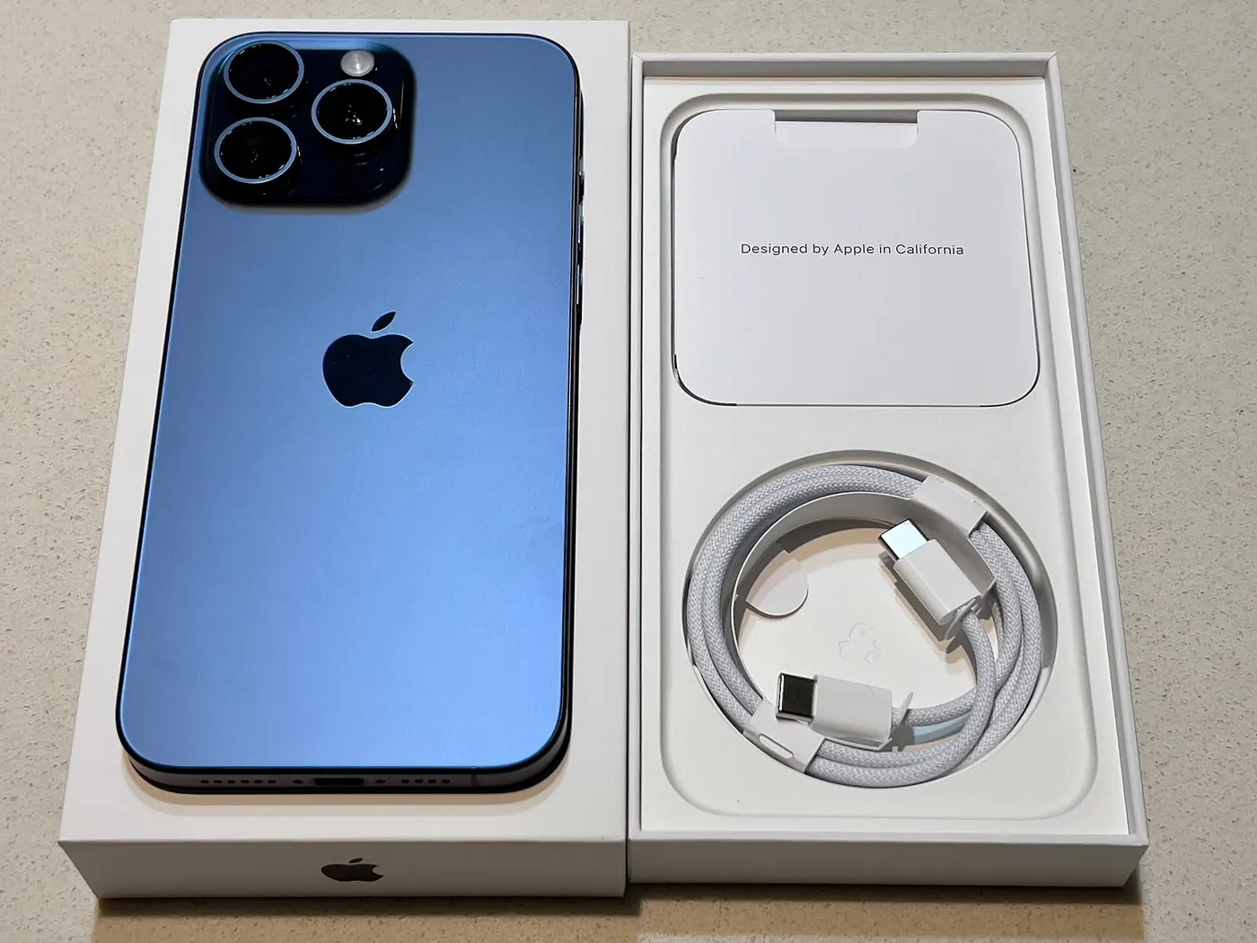 iPhone 15 Pro Max en color titanio azul junto a su caja abierta y el cable USB-C