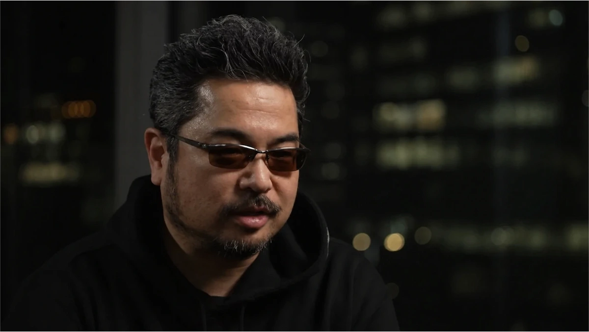 Katsuhiro Harada, con anteojos oscuros, durante una entrevista con luces de la ciudad de fondo