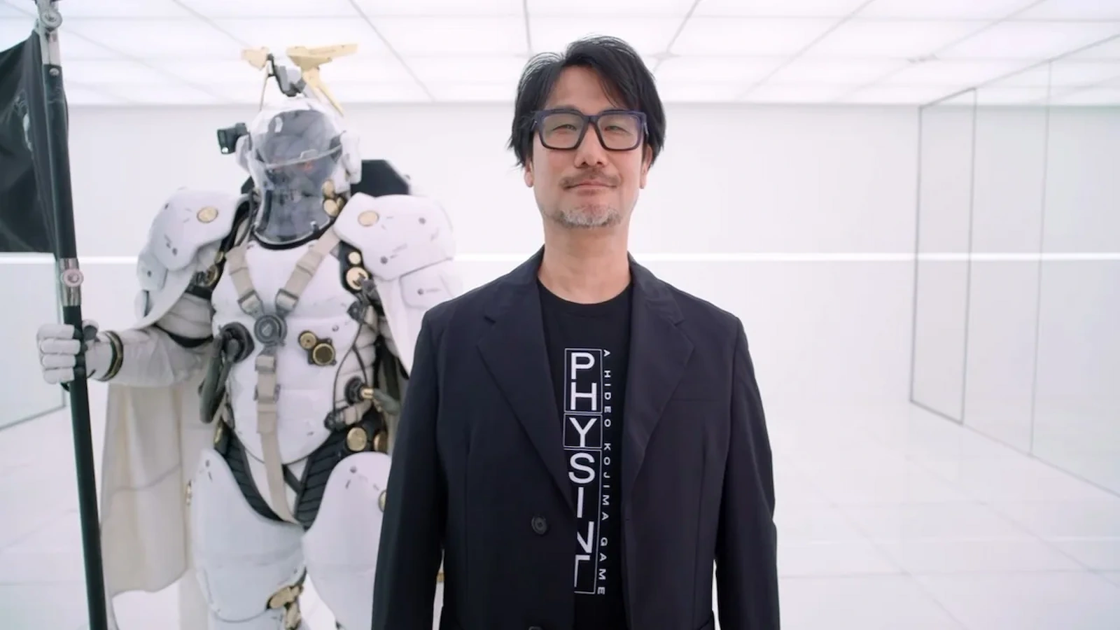 Hideo Kojima con una remera de PHYSINT, junto a una figura de Ludens, la mascota de Kojima Productions