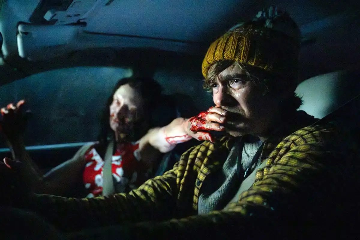 Austin Abrams dentro de un auto con una mujer ensangrentada que le tapa la boca, en Resident Evil: Noche Cero