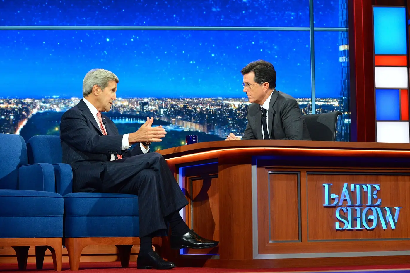 Stephen Colbert detrás del escritorio de The Late Show, con el logo del programa a la vista, durante la entrevista al secretario de Estado John Kerry