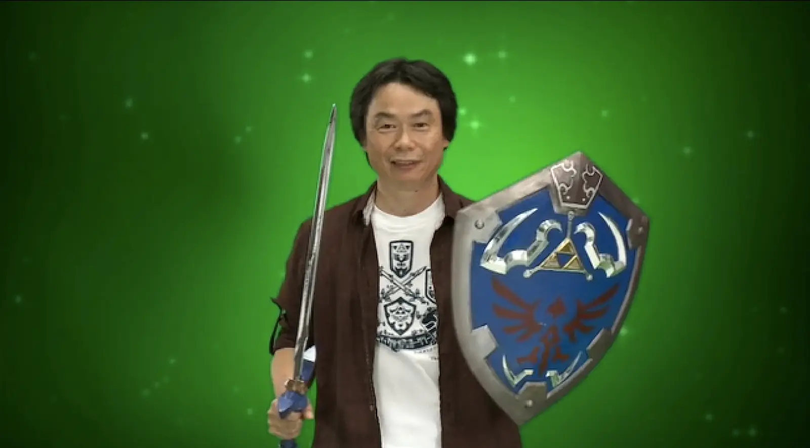 Shigeru Miyamoto con la Espada Maestra y el escudo hyliano de The Legend of Zelda frente a un fondo verde