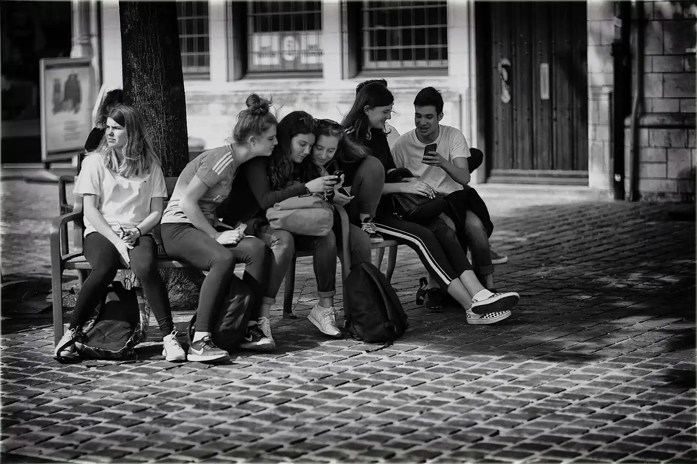 Un grupo de adolescentes sentados en un banco de la calle mira sus celulares, en blanco y negro