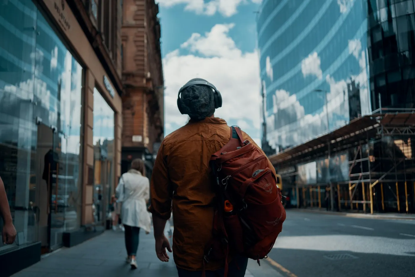 Hombre con campera naranja y auriculares inalámbricos caminando de espaldas por una calle de la ciudad