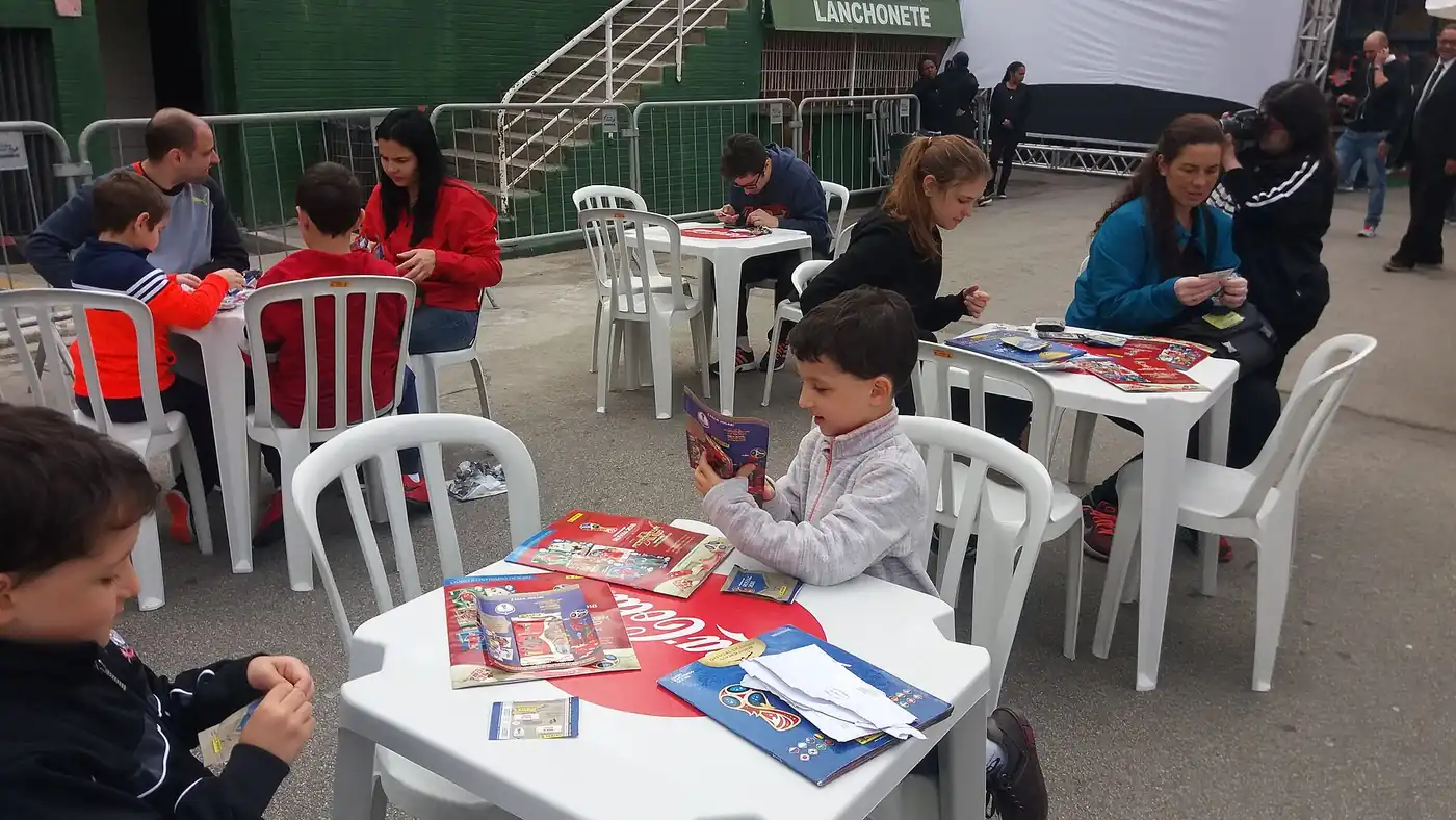 Chicos y adultos sentados en mesas al aire libre completando álbumes de figuritas de Panini
