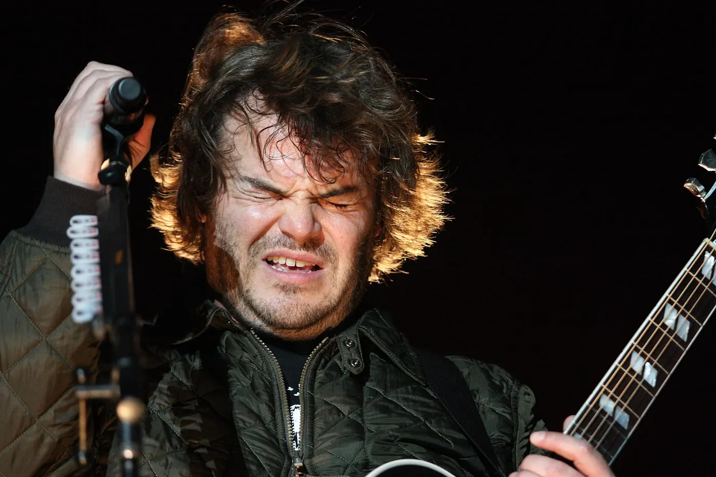 Jack Black canta con los ojos cerrados frente al micrófono y con la guitarra colgada, en un show de Tenacious D