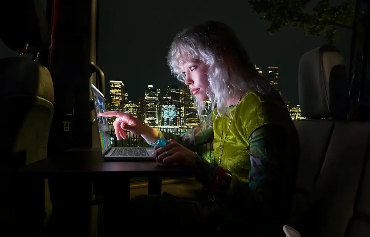 Una usuaria toca la pantalla de la MacBook Pro con PROLED XDR de noche, con una ciudad iluminada de fondo