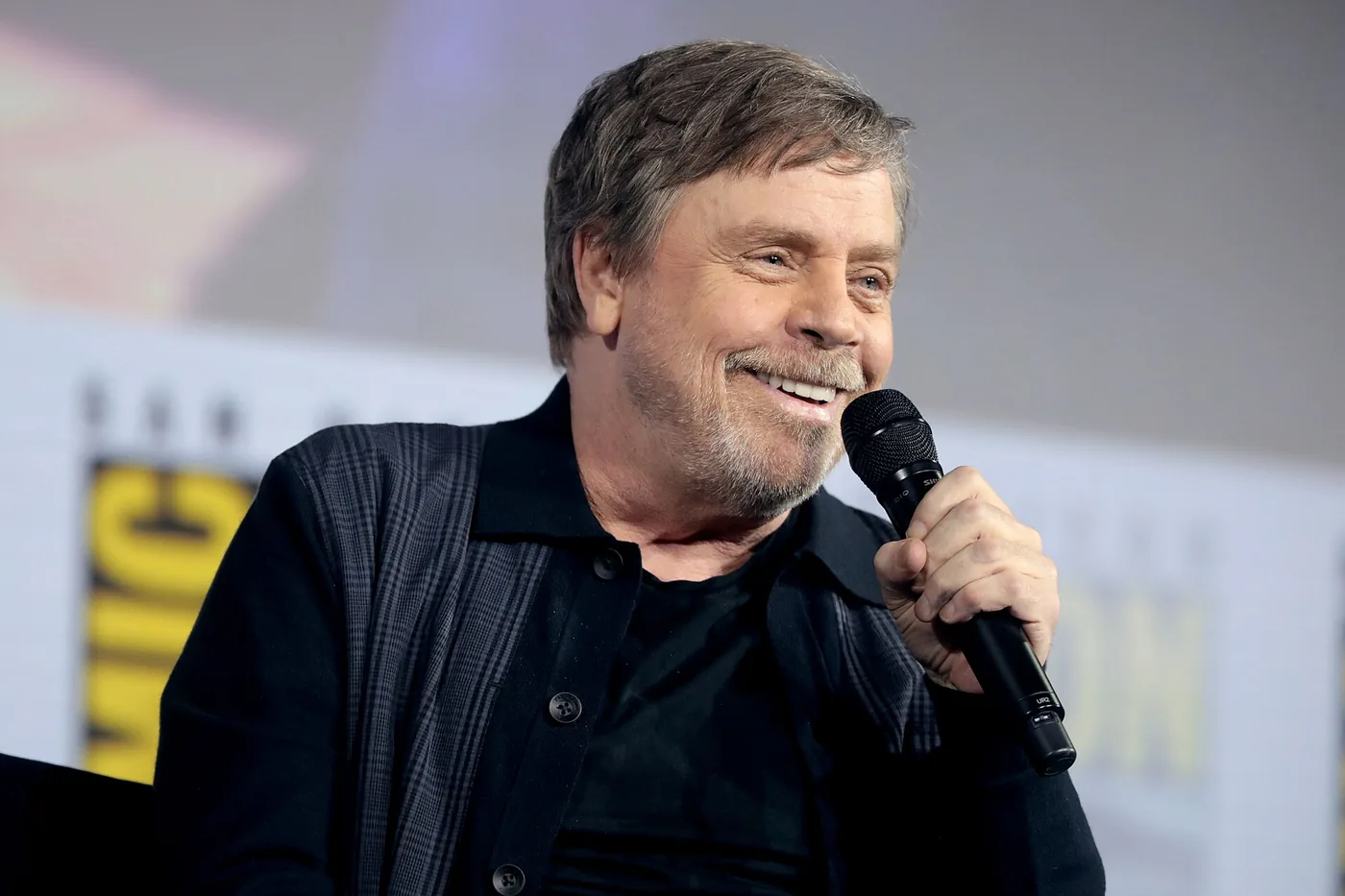 Mark Hamill sonríe con un micrófono en la mano durante un panel de la Comic-Con de San Diego