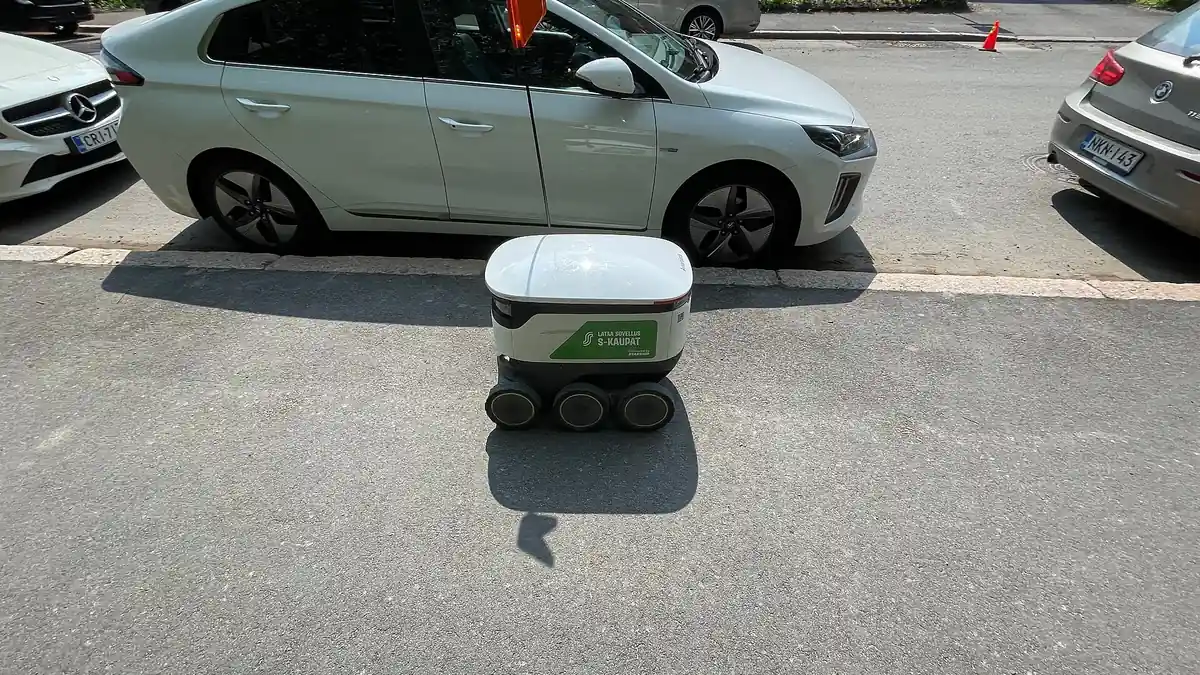 Robot de reparto autónomo de seis ruedas cruza una calle frente a autos estacionados